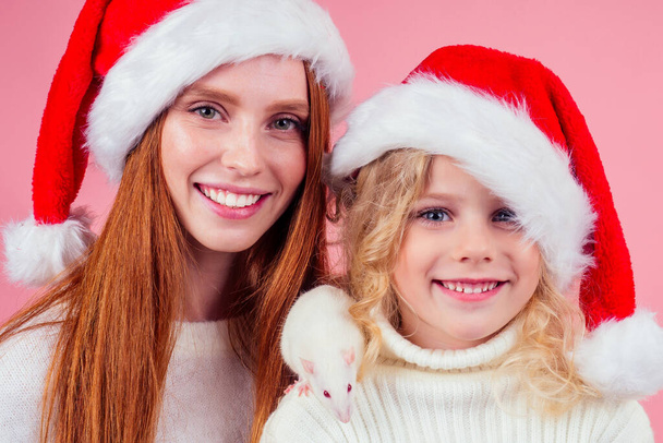 redhaired ginger woman with small little blond girl wearing santas hat holding many gift boxes with whire rat in studio pink background - Фото, зображення