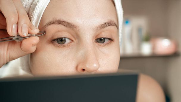 Plucking eyebrows. Girl with white towel on her head in bathroom looks in mirror and plucks her eyebrow hair with tweezers. Hygiene, beautician, makeup, face care concept. Close-up view - Fotó, kép