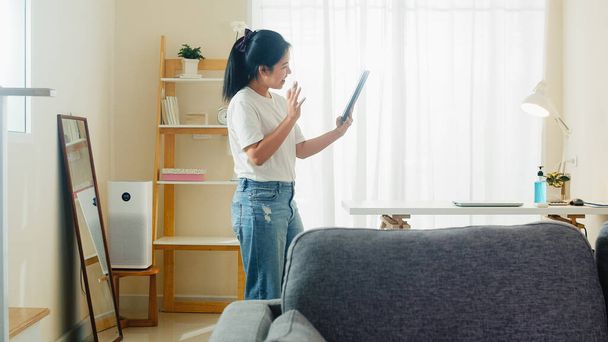Asyalı serbest çalışan kadın günlük kıyafet tablet çalışma video konferansı kullanarak evden oturma odasına müşteri çağırma sosyal uzaklık evde ve kendi kendine karantina zaman. - Fotoğraf, Görsel