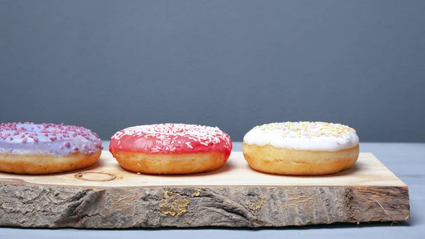 разноцветные красные, белые, фиолетовые пончики, покрытые глазурью и посыпанные пасхальным порошком на деревянный стенд на сером фоне, сладкая эко-еда
  - Фото, изображение