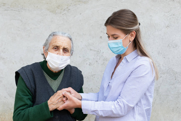 Portrait of friendly caregiver posing with elderly ill woman wearing surgical mask because of covid-19 pandemic - Фото, зображення