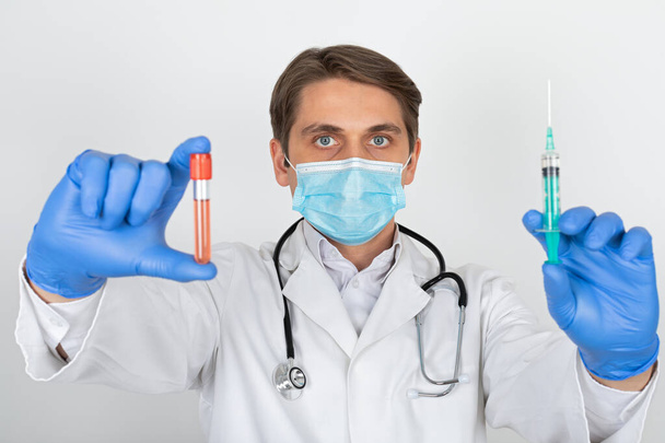 Young male doctor with surgical mask and gloves holding a blood sample and injection on white background - Фото, зображення