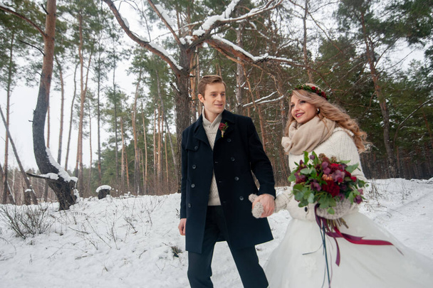 Vrolijke bruid en bruidegom in beige gebreide truien lopen in besneeuwd bos. Winterbruiloft - Foto, afbeelding
