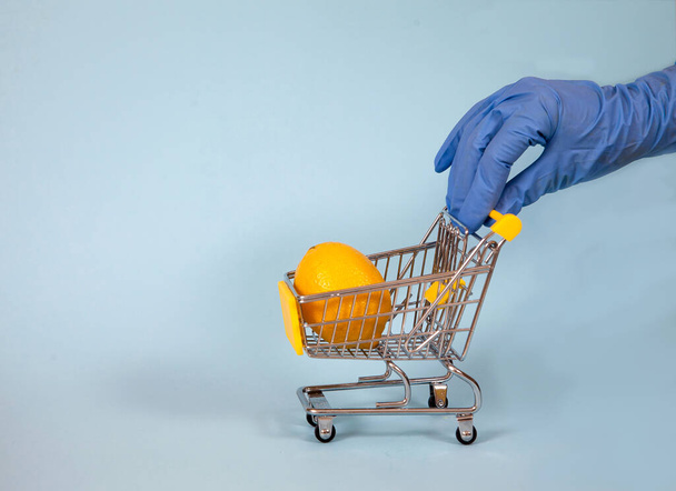 Female hand in a blue rubber glove rolls a supermarket trolley with lemon on a blue background. Protection. Hygiene. covid 19. copy spaes - Φωτογραφία, εικόνα