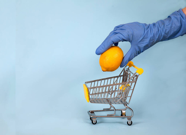 Female hand in a blue rubber glove lowers a lemon into a supermarket trolley on a blue background. Protection. Hygiene. Health. Copy spaes. covid 19. - Φωτογραφία, εικόνα