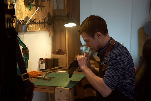 Arbeitsprozess des Ledermanns in der Werkstatt. Junge Handwerker bearbeiten Naturleder mit Bastelwerkzeugen. Arbeiten mit echtem Leder. Ein kleiner Handwerksbetrieb. Das Foto kann für Handarbeiten, Kunsthandwerk, die Herstellung von Lederprodukten verwendet werden - Foto, Bild