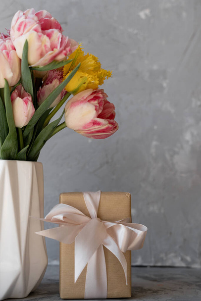 Bunte Tulpen in einer weißen Vase auf dem Tisch vor hellgrauem Hintergrund, Schachtel mit Geschenk. Kraft Papier und Bogen. Platz für Text. Muttertag - Foto, Bild