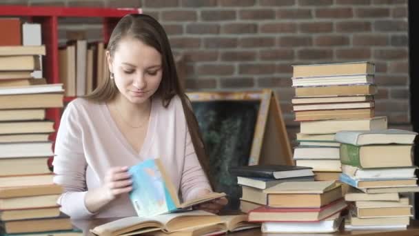 beautiful young girl is sitting in room home table and reading book. student studies home. Exam preparation. woman reads textbook among a lot of books. home education. The concept of modern education. - Footage, Video