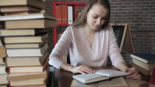 φοιτητής διαβάζει βιβλία. όμορφη νεαρή κοπέλα κάθεται σε ένα δωμάτιο στο σπίτι σε ένα τραπέζι και ξεφυλλίζοντας τις σελίδες ενός βιβλίου. Μια γυναίκα διαβάζει ένα βιβλίο ανάμεσα σε πολλά βιβλία. Προετοιμασία εξετάσεων. - Πλάνα, βίντεο