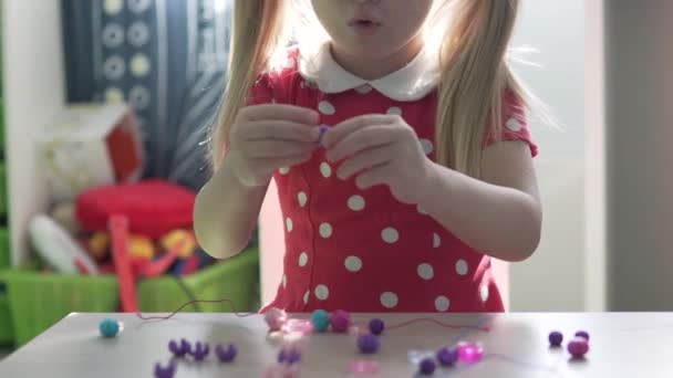 Ein kleines Mädchen fertigt zu Hause am Tisch ein Armband aus Perlen - Filmmaterial, Video