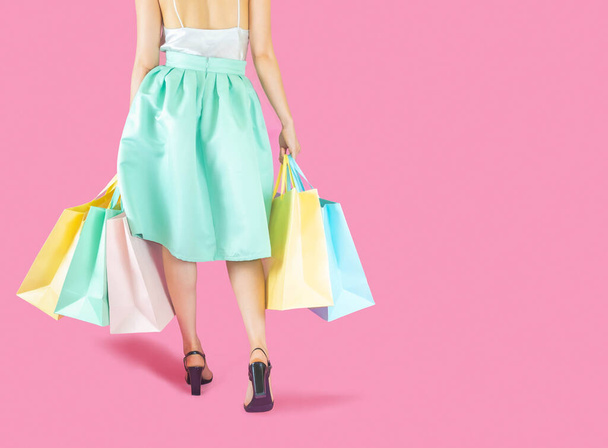The back of woman low body part wore blue skirt and black high heels. Carrying a shopping bag in many pastel colors on pink background selective focus - Photo, image