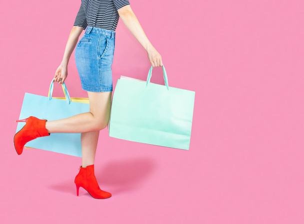 The female body part wore pink skirt jean and red boots. Carrying a shopping bag in many pastel colors on pink background selective focus,shopping concept - Zdjęcie, obraz
