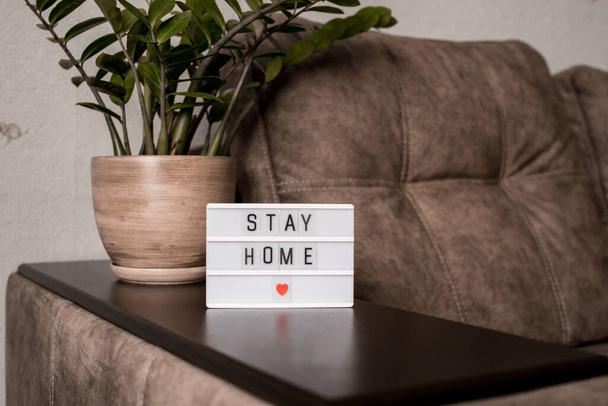 Uma caixa de luz com a inscrição ficar em casa fica na parte de trás de um sofá com uma flor do quarto. Conceito
 - Foto, Imagem