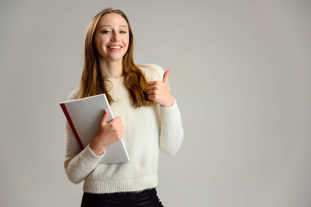 Studio photo of a blonde student girl on a white background in a white sweater with a folder in her hands - Fotoğraf, Görsel