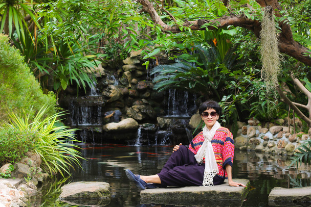 Senior woman wear northern red shirt and trouser with white scarf sitting on stone with waterfall and tree background. - Фото, изображение