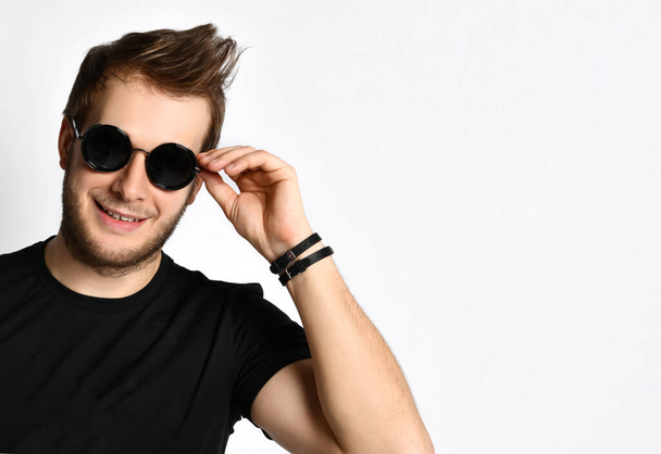 Hipster-Typ in schwarzem T-Shirt mit Print, Armbänder. Er lächelt und justiert seine Sonnenbrille, posiert isoliert auf weiß. Nahaufnahme - Foto, Bild