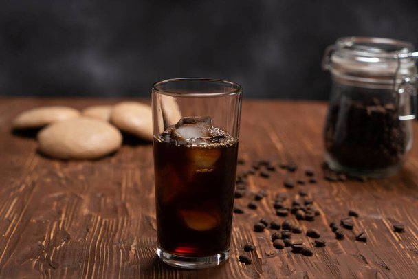 Glass of cold brew iced coffee on brown wood table with coffee beans and cookies on black background - Foto, immagini