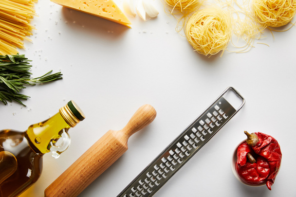 Bovenaanzicht van deegroller, fles olijfolie, rasp, pasta en ingrediënten op witte achtergrond  - Foto, afbeelding