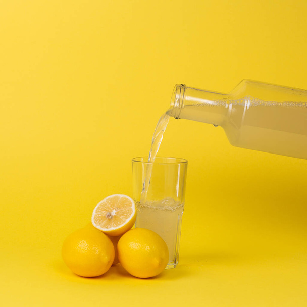 Zitronensaft wird mit einer Glasflasche in ein Glas gegossen. Ganze und geschnittene Zitronen liegen in der Nähe. - Foto, Bild