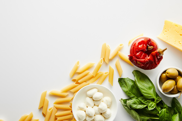 Top view of pasta, basil leaves, cheese and bowls with olives, marinated chili peppers and mozzarella on white background  - Photo, Image