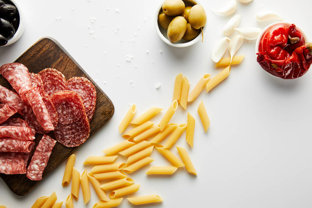 Bovenaanzicht van pasta, zeezout, knoflook, vleesschotel en schalen met olijven en gemarineerde chili pepers op witte achtergrond  - Foto, afbeelding