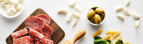 Panoramic orientation of meat platter, rolling pin, pasta, garlic and bowls with olives and mozzarella on white background - Photo, Image