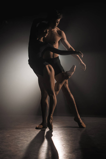 Beautiful young acrobats or gymnasts on floodlights background. Professional ballet couple dancing in spotlights smoke on big stage. Emotional duet performing choreographic art - Foto, imagen
