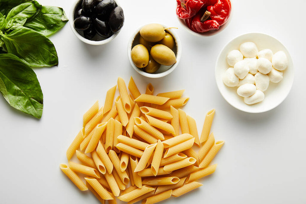 Draufsicht auf Pasta in der Nähe von Basilikumblättern und Schalen mit Zutaten auf weißem Hintergrund  - Foto, Bild