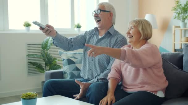 Joyful old lady watching TV with husband laughing having fun enjoying movie at home - Materiał filmowy, wideo