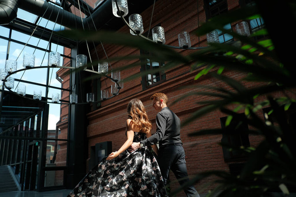 schönes Paar - ein Mädchen in einem prächtigen Kleid und ein Mann in einem schwarzen Hemd und einer schwarzen Hose befinden sich in einem Gebäude mit einem wunderschönen Interieur - Foto, Bild