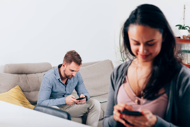 άνδρας στο ζευγάρι γυναικών που κάθεται χωριστά στο σπίτι κοιτάζοντας το κινητό τους τηλέφωνο - τηλέφωνο εθισμένος, κλείδωμα και καραντίνα τρόπο ζωής - Φωτογραφία, εικόνα