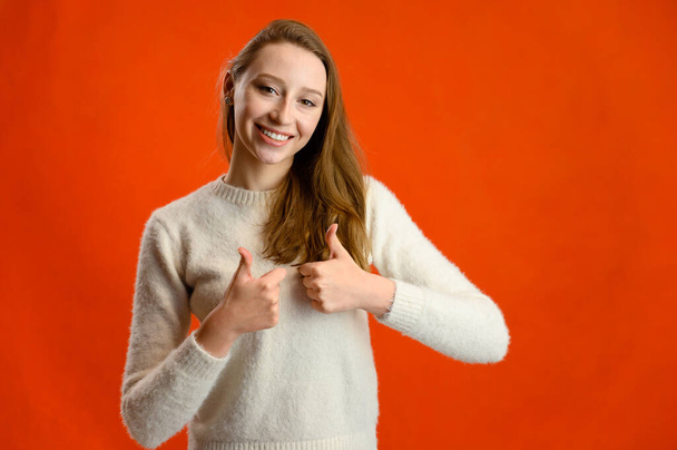 Model talking showing happy emotions showing fingers. Studio photo of a cute caucasian girl in a white sweater standing in front of the camera on a red background. - Foto, imagen