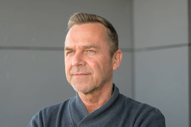 Attractive serious trendy middle-aged man in grey sweater standing in the corner angle of a room looking thoughtfully at the camera with an intense stare - Photo, Image
