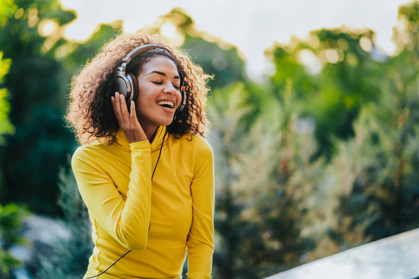 African teenager girl listens to music with headphones in park. Cute woman in yellow, dancing with head, curly hair. Student, freedom, music concept. - Foto, afbeelding