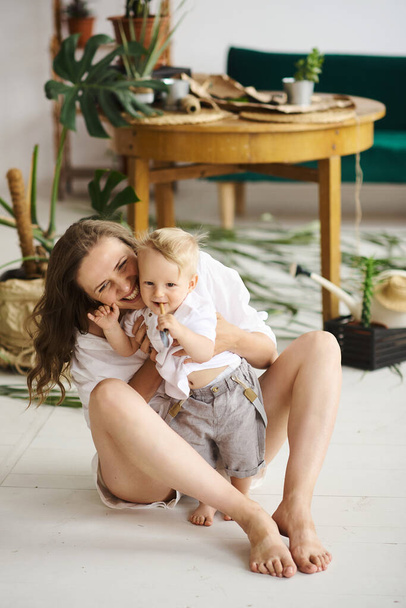 Une jeune belle maman jouant avec son bébé sur le sol près de la table en bois avec des plantes et un canapé vert
 - Photo, image