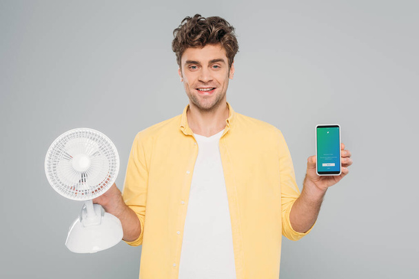 KYIV, UKRAINE - MARCH 27, 2020: front view of man smiling, looking at camera and showing desk fan and smartphone with twitter app isolated on grey - Photo, Image