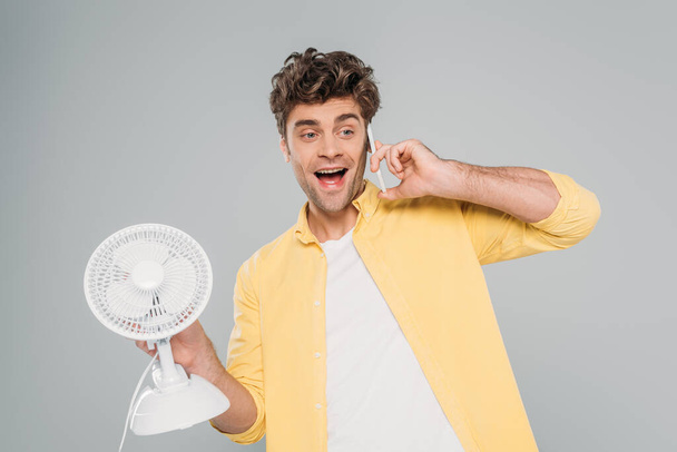 Front view of excited man holding desk fan and talking on smartphone isolated on grey - Photo, Image