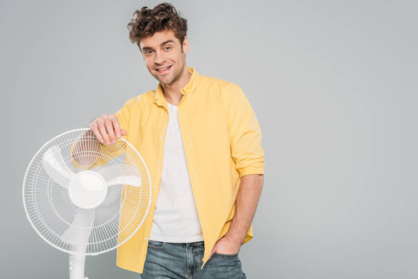 Front view of man with hand in pocket smiling and looking at camera near electric fan isolated on grey - Photo, Image