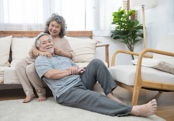Portret starszej pary azjatyckiej szczęśliwej razem patrzeć w kamerę w domu.Dziadek siedzi na podłodze opierając się na babci siedzieć w autokarze w salonie w domu mają wolny czas w stylu życia emerytalnego. - Zdjęcie, obraz