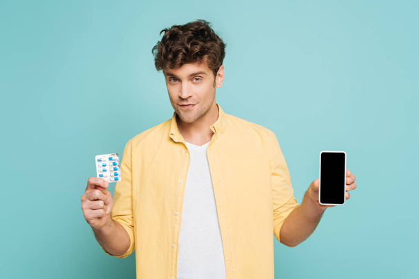 Vorderansicht eines lächelnden Mannes, der in die Kamera blickt und Smartphone und Blisterpackung mit Tabletten auf blauem Grund zeigt - Foto, Bild
