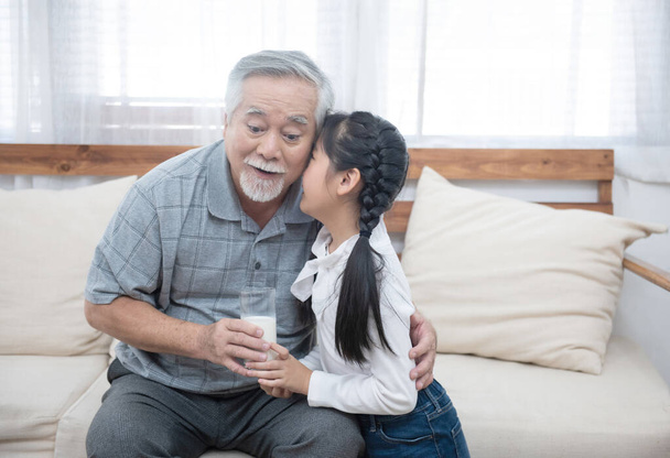 Ευτυχισμένος Ασιάτης ηλικιωμένος παππούς έχουν εγγόνι φροντίζει και να φροντίσει με δίνοντας γάλα και φιλί στο μάγουλο, ενώ κάθεται στον καναπέ στο σπίτι, συνταξιοδότηση έννοια του τρόπου ζωής της υγείας. - Φωτογραφία, εικόνα