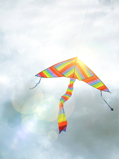 Bunte Kinderdrachen fliegen unter einem wolkenverhangenen Himmel, durch den Sonnenstrahlen lugen. - Foto, Bild
