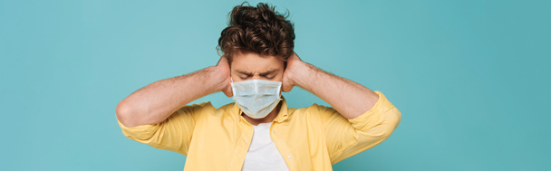 Front view of man in medical mask with closed eyes covering ears isolated on blue, panoramic shot - Photo, Image