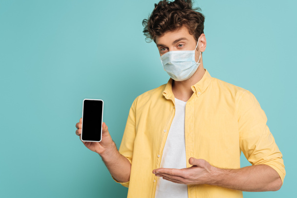 Mężczyzna w masce medycznej wskazuje na odizolowanego na niebiesko smartfona - Zdjęcie, obraz
