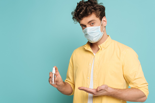 Man in medical mask pointing at bottle of hand sanitizer isolated on blue - Foto, afbeelding