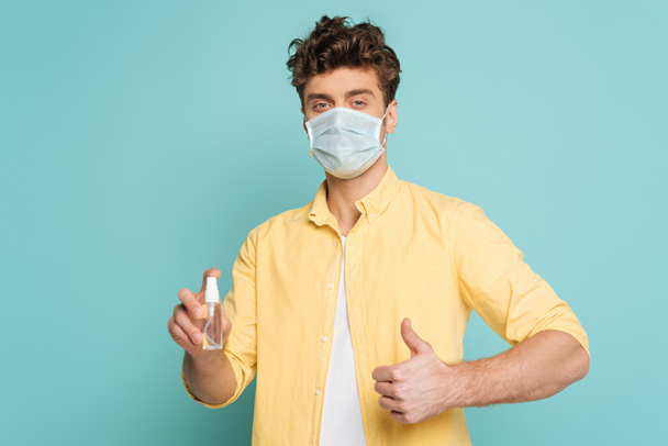 Mann in medizinischer Maske hält Flasche mit Händedesinfektionsmittel und zeigt wie ein Zeichen isoliert auf blauem Grund - Foto, Bild