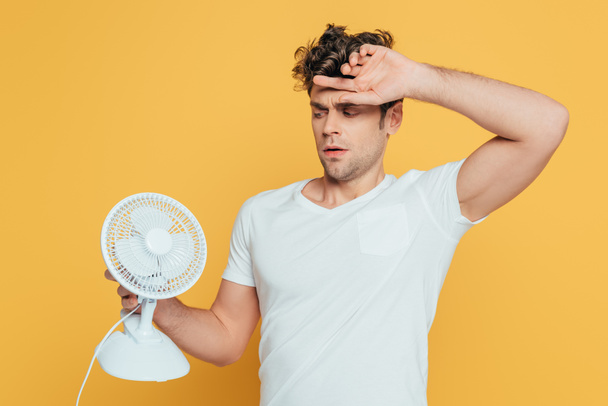 Confused man with raised hand looking at desk fan isolated on yellow - Foto, imagen