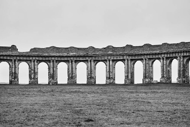 antiguas ruinas de antiguas paredes arqueadas de ladrillo en un prado verde vacío y sobre el fondo del cielo vacío y limpio de tono monocromo
 - Foto, imagen