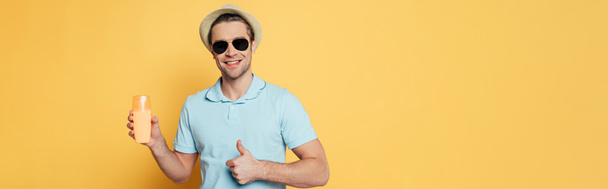 Front view of man in hat and sunglasses with bottle of sunscreen smiling and showing like sign isolated on yellow, panoramic shot - Foto, immagini
