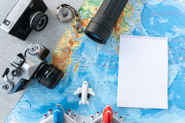 Flache Glasscheibe, eine alte seltene Filmkamera, ein Kompass, ein weißes Notizbuch für Reisenotizen, drei kleine Flugzeugspielzeuge auf geografischer Landkarte. Reisekonzept, Urlaubsplanung - Foto, Bild